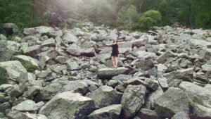 A person stands upon large rocks covering the ground with arms opened wide.