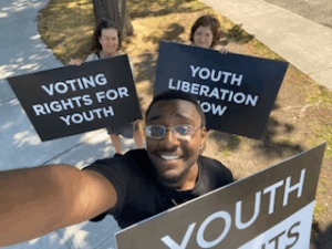 Eric Kennedy, Imogen Gannon, and Susan Milton holding large signs reading Youth Rights, Youth Liberation Now and Voting Rights For Youth respectively, in all caps.