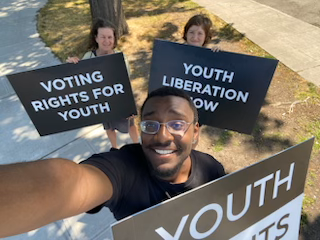 Eric Kennedy, Imogen Gannon, and Susan Milton holding large signs reading Youth Rights, Youth Liberation Now and Voting Rights For Youth respectively, in all caps.